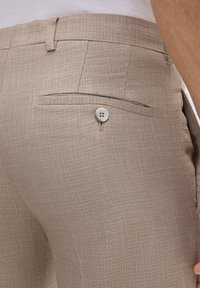 Beige Hosen aus strukturiertem Stoff, mit einer Gesäßtasche und runden Knopfdetails. Einfache Webstruktur, schlankes Design.