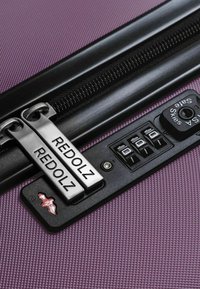 Valigia viola con texture, cerniera nera e lucchetto a combinazione. Presenta tiranti della cerniera argentati etichettati "REDOLZ". Design compatto con hardware sicuro.
