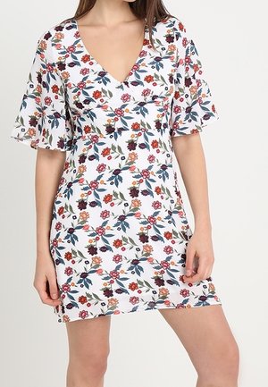 Femme portant une courte robe blanche à motifs floraux roses, rouges et oranges avec des feuilles vertes, avec un col en V et des manches courtes évasées.