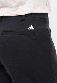 Μαύρο παντελόνι με ένα μικρό λευκό τριγωνικό λογότυπο κοντά στην πίσω τσέπη, φορεμένο με λευκό πουκάμισο που έχει πορτοκαλί σχέδιο.