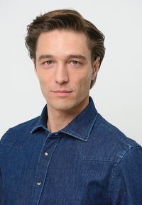 Camicia in denim blu scuro con collo a punta, taschino sul petto e due bottoni frontali. Tessuto liscio con impunture visibili.