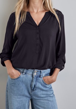 Frau trägt eine schwarze, langärmelige, geknöpfte Bluse, die in eine hoch taillierte hellblaue Jeans gesteckt ist, mit den Händen in den Taschen.