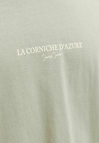Tissu vert clair avec texte imprimé blanc "LA CORNICHE D'AZURE" et une petite signature en cursive en dessous.