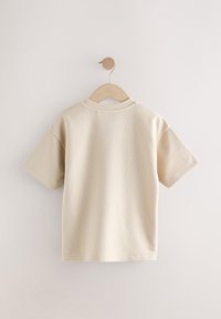 Camicia beige a maniche corte in tessuto strutturato con motivo a griglia. Presenta una scollatura tonda ed è appesa a una gruccia di legno su uno sfondo chiaro.