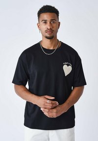 Camiseta de algodón negra con un parche en forma de corazón color crema en el lado izquierdo del pecho. Mangas cortas y un corte relajado. Detalle de costura simple.