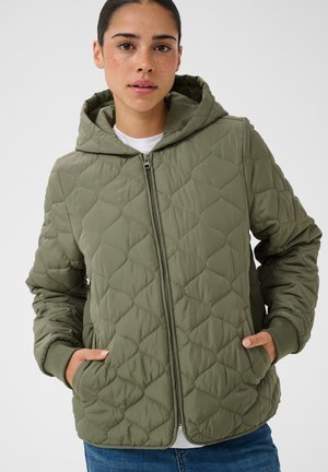 Jeune femme portant une veste matelassée verte à capuche zippée, les mains dans les poches, associée à une chemise blanche et un jean bleu.