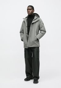 Šedá nepremokavá bunda s kapucňou, so zipsom a gombíkovým zapínaním, skombinovaná s čiernymi cargo nohavicami a textúrovanými čiernymi topánkami. Minimalistický dizajn.