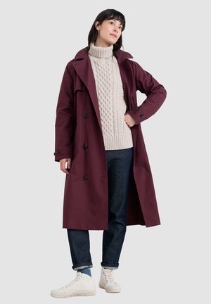 Bordeaux trenchcoat met een dubbelzijdig ontwerp, zwarte knopen, gecombineerd met een crèmekleurige kabelgebreide trui en donkere spijkerbroek, aangevuld met witte sneakers.