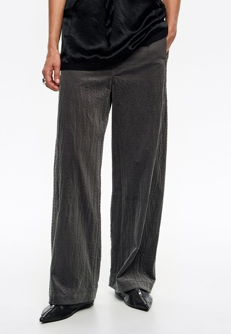 Pantalones de pierna ancha gris, hechos de un tejido texturizado con acanalado vertical, que presentan un ajuste relajado y se combinan con zapatos negros de punta.