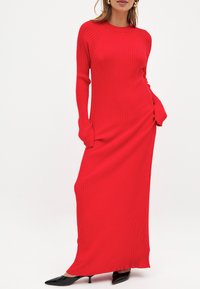 Sarkans rīvēts maxi kleita ar garām piedurknēm, pieguļošu siluetu, sānu rušinga detaļu un mīkstu tekstūru. Valkāta ar melnajām norādītajām kurpēm.