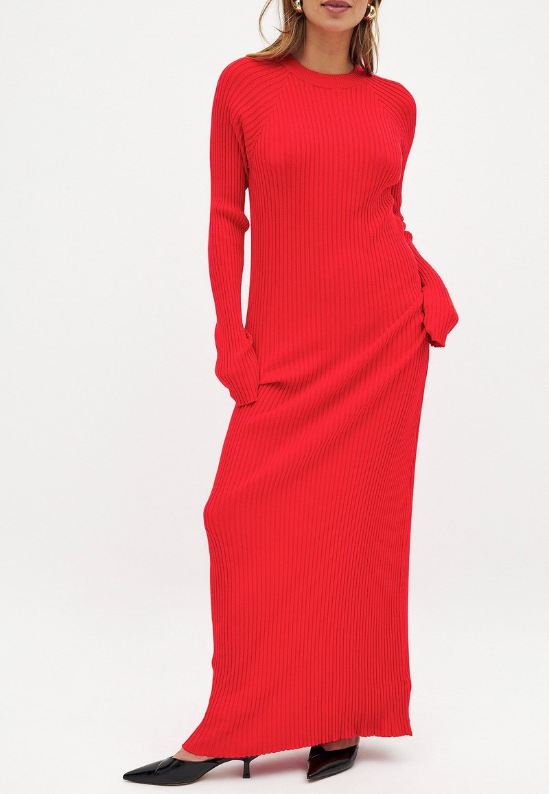 Sarkans rīvēts maxi kleita ar garām piedurknēm, pieguļošu siluetu, sānu rušinga detaļu un mīkstu tekstūru. Valkāta ar melnajām norādītajām kurpēm.