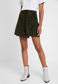 Olivgrön kordkjol med ett brett bälte som har en cirkulär guldbuckle, kombinerad med vita ankelboots i ormskinnsmönster.