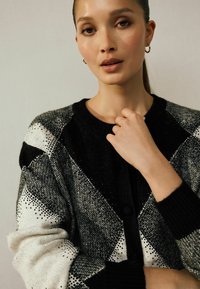 Svetr s argyle vzorem v černobílé, strukturovaný pletený materiál, úzké manžety, zapínání na knoflíky, viditelné spečky metalických akcentů.