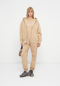 Felpa beige e pantaloni jogger coordinati, realizzati in tessuto morbido, con un logo stampato sulla maglia. Borsa nera e scarpe sportive grigie.