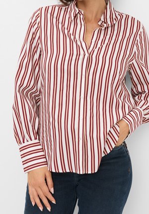 Frau trägt ein langärmeliges, zum Knöpfen geeignetes Hemd mit vertikalen rot-weißen Streifen, kombiniert mit dunkelblauen Jeans.
