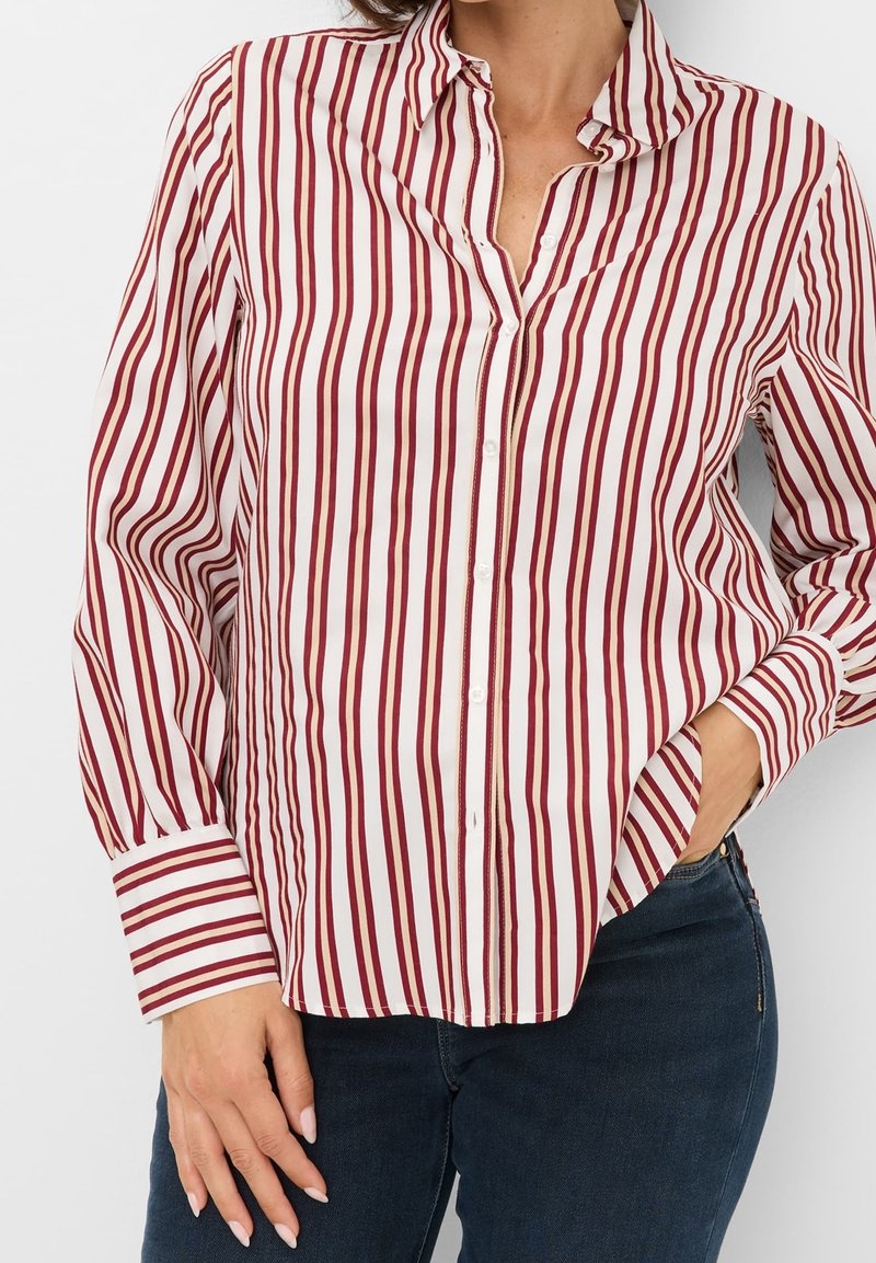 Femme portant une chemise à manches longues boutonnée avec des rayures verticales rouges et blanches, associée à un jean bleu foncé.