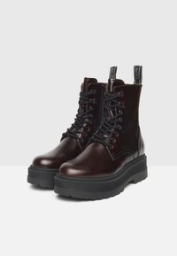 Bottes chelsea en cuir marron avec une épaisse semelle noire, bout rond, à lacets à l'avant et languettes sur le col. Fini lisse, accents minimaux.