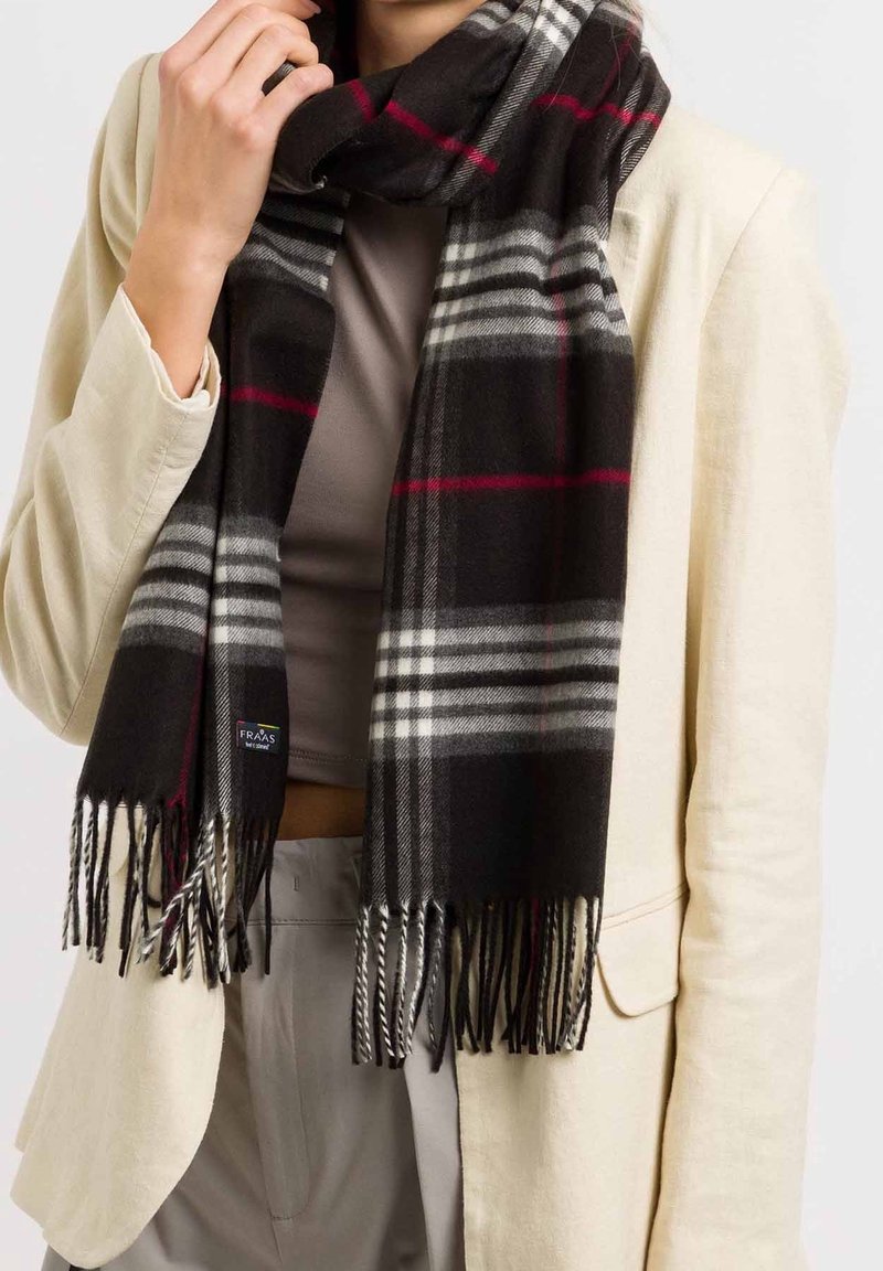 Personne portant un blazer beige et un haut taupe, tenant une écharpe à carreaux noire avec des rayures blanches et rouges et des extrémités à franges autour du cou.