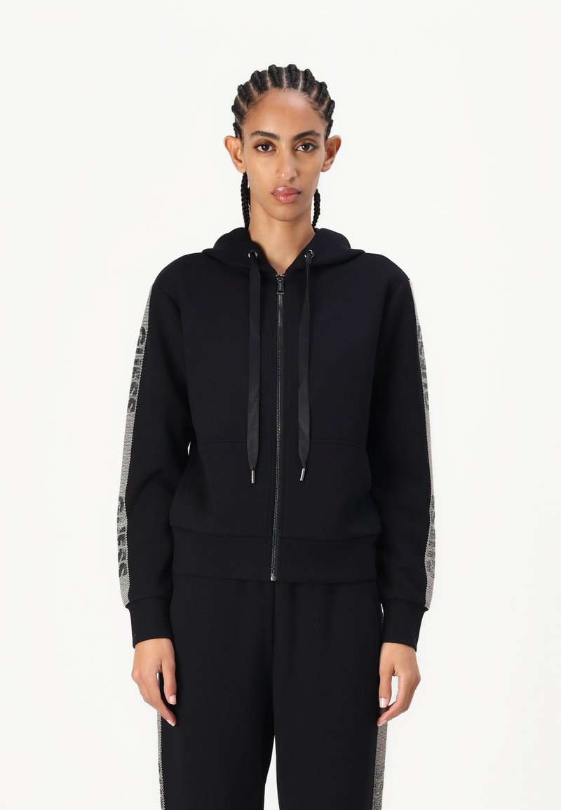Sudadera negra con cremallera y bolsillo frontal, con acentos texturizados en las mangas y cordones. Hecha de una tela suave con un acabado liso.