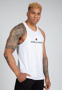 Fehér, ujjatlan trikó "GORILLA WEAR" logóval, laza szabással és puha anyaggal, fekete nadrággal és több tetoválással párosítva.