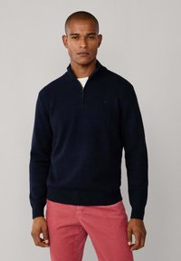 Marinefarbener Strickpullover mit Reißverschlusskragen, gerippten Bündchen und Saum. Getragen über einem weißen Hemd, kombiniert mit hellrosa Hosen.