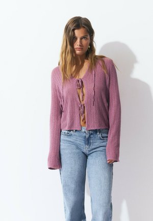Femme portant un cardigan en tricot violet clair avec deux noeuds à l'avant et un jean taille haute bleu clair contre un mur blanc.