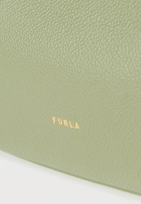 Επιφάνεια από δέρμα ανοιχτού πράσινου χρώματος με υφή, με μικρό χρυσό κείμενο "FURLA" ανάγλυφο κοντά στο κέντρο του κάτω μέρους.