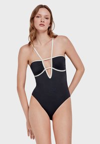 Maillot de bain noir une pièce avec un décolleté plongeant, doté de bretelles crème et de tissu froncé au niveau de la poitrine. Texture lisse et soyeuse.