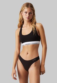 Bralette negra con banda ancha de logotipo blanco y escote en forma de scoop, combinada con bragas de bikini negras a juego que presentan un sutil detalle de logotipo.