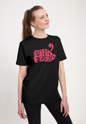Ung kvinde med langt hestehale iført sort Pink Floyd t-shirt kigger op og smiler mod ensfarvet baggrund.