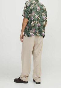 Man i lösa beige byxor, mörkbruna skor och en grön kortärmad skjorta med rosa och vita blommönster, stående mot en enhetlig bakgrund.