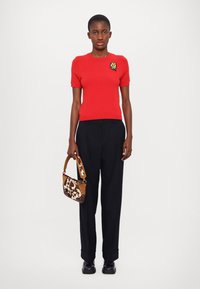 Maglietta rossa a maniche corte in maglia con un colorato patch ricamato, abbinata a pantaloni neri a vita alta e una borsa marrone a pois.
