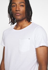 Άνδρας με μεσαίου μήκους κοτσίδες dreadlocks, που φοράει απλό λευκό κοντομάνικο μπλουζάκι με μικρή τσέπη στο στήθος, σε λευκό φόντο.