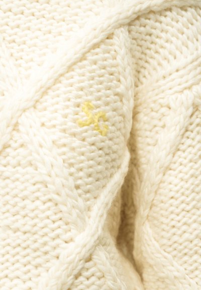 Κρεμ πλεκτό ύφασμα με σχεδιασμούς σε σχήμα καλωδίου και μικρή κίτρινη κεντημένη λεπτομέρεια κοντά στο κέντρο.