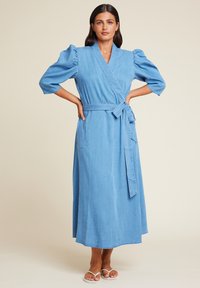 Lys blå denim omslagskjole med korte puffermer, midjebånd og en lang, flytende skjørt. Glatt tekstur og enkelt design.