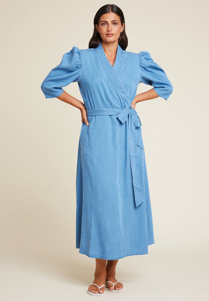 Lys blå denim omslagskjole med korte puffermer, midjebånd og en lang, flytende skjørt. Glatt tekstur og enkelt design.