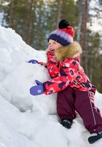 Jasno kolorowa kurtka narciarska w kwiatowy wzór z futrzanym wykończeniem, w połączeniu z bordowymi spodniami narciarskimi i purpurowymi rękawiczkami, noszona przez dziecko na śniegu.