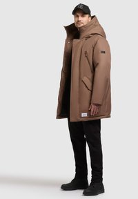 Brauner Puffercoat mit hohem Kragen, Reißverschlussfront und seitlichen Taschen. Getragen über schwarzen Hosen und schwarzen Schuhen, ergänzt durch eine Mütze.