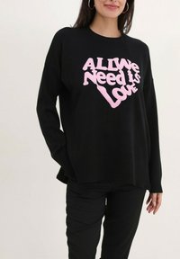 Pull noir à manches longues, avec une inscription en relief rose qui dit "Tout ce dont nous avons besoin, c'est d'amour", avec une coupe ample et décontractée.