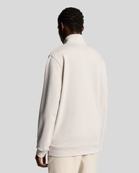 Personne avec des cheveux tressés portant un sweat-shirt beige clair uni et un pantalon assorti, se tenant de dos contre un fond gris.