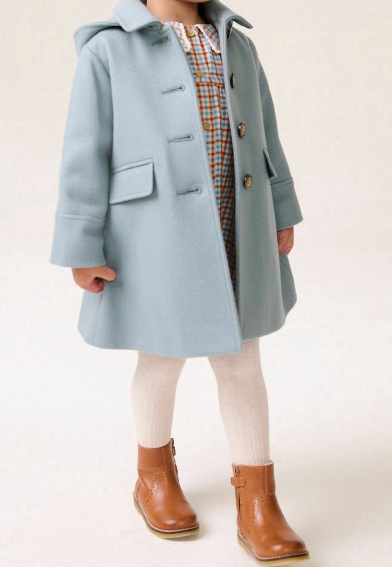 Enfant portant un manteau à capuche bleu clair par-dessus une robe à carreaux, des collants crème et des bottines en cuir marron sur un fond uni.