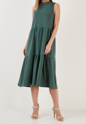 Frau trägt ein ärmelloses, dunkelgrünes Stufenkleid in Midilänge und beige hochhackige Sandalen, steht vor einem schlichten weißen Hintergrund.