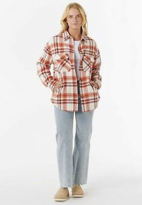 Chemise en flanelle à carreaux rouge et crème avec deux poches poitrine, fermetures à boutons, et coupe décontractée ; portée par-dessus un haut blanc et associée à un jean clair.