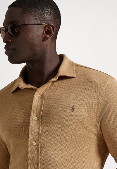 Vyras dėvintis akinius nuo saulės su vėžlio kiauto raštu ir smėlio spalvos herringbone rašto marškinius su mažyčiu siuvinėtu logotipu ant krūtinės, žvelgiantis į kairę.