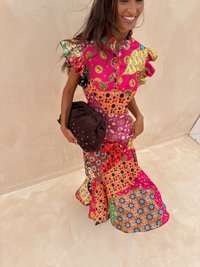 Kleurrijke patchwork jurk met gerimpelde mouwen, met levendige patronen in roze, oranje en groen. Houdt een bruine tas met gekleurde accenten vast.