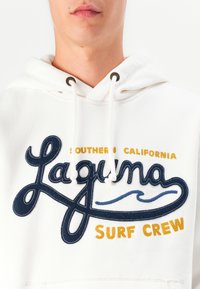 Felpa bianca con cappuccio realizzata in tessuto morbido. Presenta la scritta "Laguna Surf Crew" ricamata in blu navy e giallo, con cappuccio regolabile e occhielli in metallo.