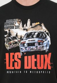 Čierne tričko s grafickou potlačou dvoch pretekárskych áut a panorámou mesta. Text znie "36 hodinový pretek Montana do New York City" a "LES DEUX."
