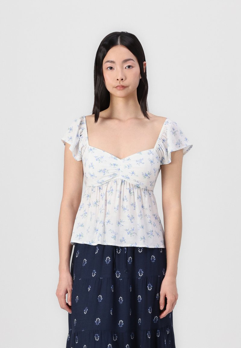 Blusa a motivi floreali in crema chiaro con fiori blu. Presenta un collo quadrato e maniche a volant. Tessuto leggero e fluido.
