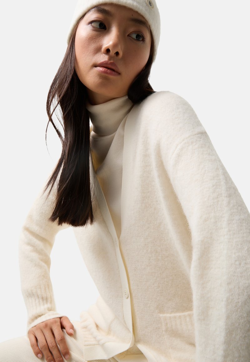 Cremfarbener Cardigan aus weichem, strukturiertem Stoff mit Knopfleiste, gerippten Bündchen und einer Tasche, über einem passenden Rollkragenpullover getragen.