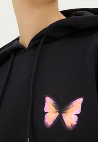 Sudadera negra con bolsillo frontal, que presenta un estampado de mariposa multicolor en rosa y naranja en el lado izquierdo. Material de algodón suave.